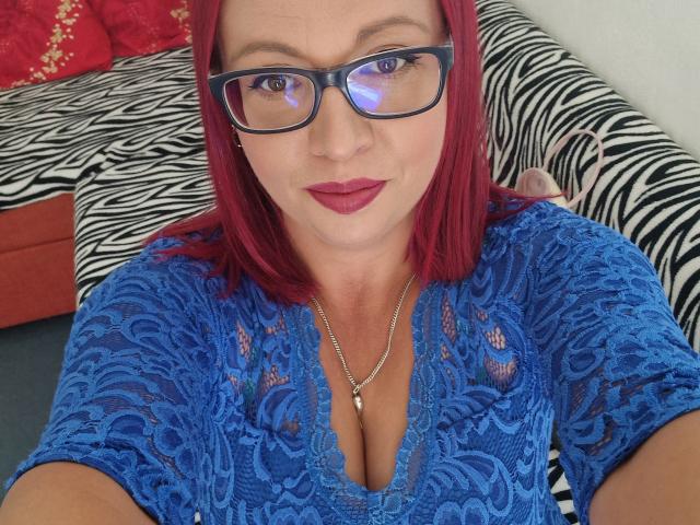 LadyHellena with red hair, blue lace top, showing her big tits, wearing glasses, red lipstick, silver necklace, sitting on zebra-patterned couch, smiling at camera.