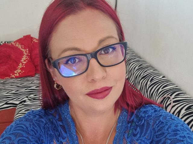 LadyHellena with red hair, blue lace top, black glasses, red lipstick, and small earrings, looking at camera, showing her tits slightly, sitting on zebra-patterned couch.