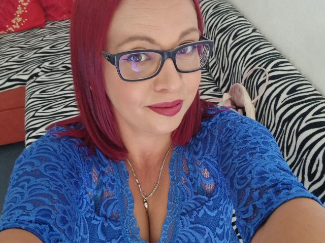 LadyHellena with red hair, glasses, blue lace top showing cleavage, wearing red lipstick, necklace with small pendant, sitting on zebra-patterned couch, holding pink high heels.