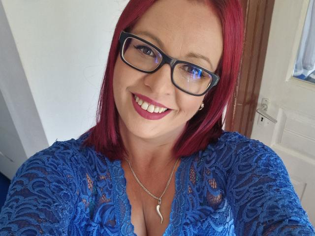 LadyHellena with red hair, black glasses, blue lace top, and a necklace showing cleavage, smiling at the camera. Boobs visible under lace. Necklace has a crescent moon pendant.