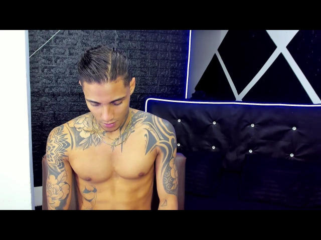 JarrodHaywardSex, shirtless, with tattoos covering his chest and arms, has dark slicked-back hair, and is looking down with a slight smirk. His ass is not visible.