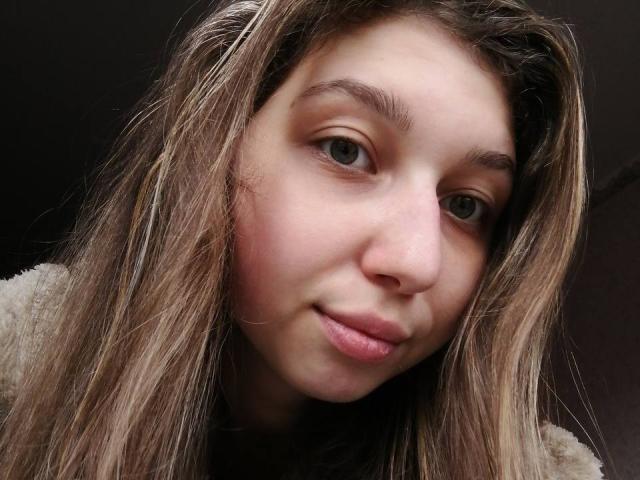 Sapfira's face, brown eyes, and light brown, wavy hair. She's smiling with pink, slightly parted lips. Her fair skin has a gentle pink blush. She's wearing a beige, fuzzy sweater.