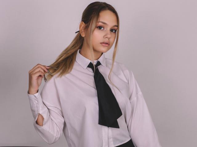 AgnesBelle has light skin, brown eyes, and blonde hair in a ponytail, wearing a white shirt and black tie, holding glasses in her right hand, looking at the camera.