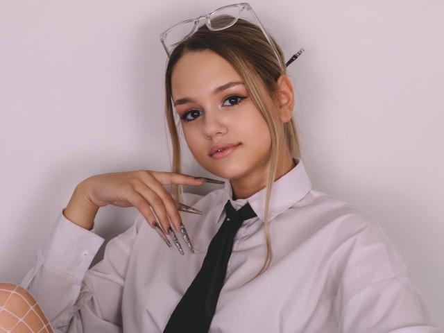 AgnesBelle with blonde hair, glasses on head, white shirt, black tie, and long nails, smiling, hand near face, showing off her tits, against a plain background.