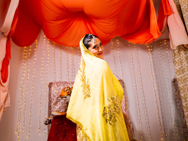 KalpanaAisha with dark hair, wearing a yellow saree with gold embroidery, looking over her shoulder, standing in front of a red draped ceiling and orange fabric.