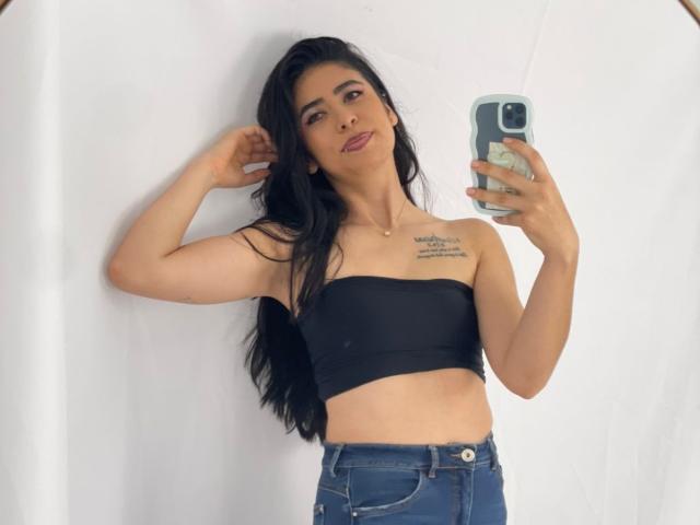 MiluSky with long black hair, wearing a black strapless top, blue jeans, holding a phone, tattoos on right shoulder, smiling, standing against a white wall.