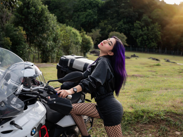 JuliaJobs mit lila Haaren, einem schwarzen Pullover und Fischernetzstrümpfen, die an einem weißen Motorrad lehnt, mit geschlossenen Augen und geneigtem Kopf, einem schwarzen Rock und einem schwarzen Rucksack, in einem grasbewachsenen Feld..