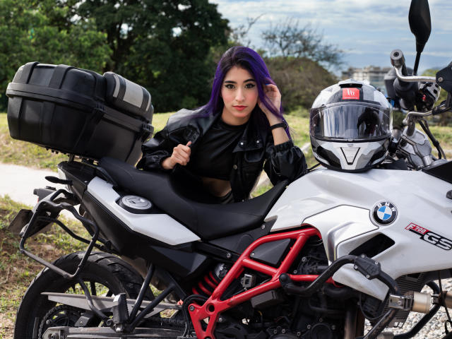 JuliaJobs with purple hair, black leather jacket, and black top, leaning on a white BMW motorcycle with red frame, black saddle bag, and white helmet.