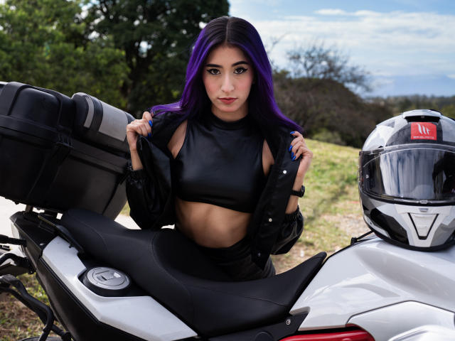 JuliaJobs with purple hair, wearing a black leather crop top, holding black jacket straps, standing in front of a white motorcycle with a black seat and helmet.