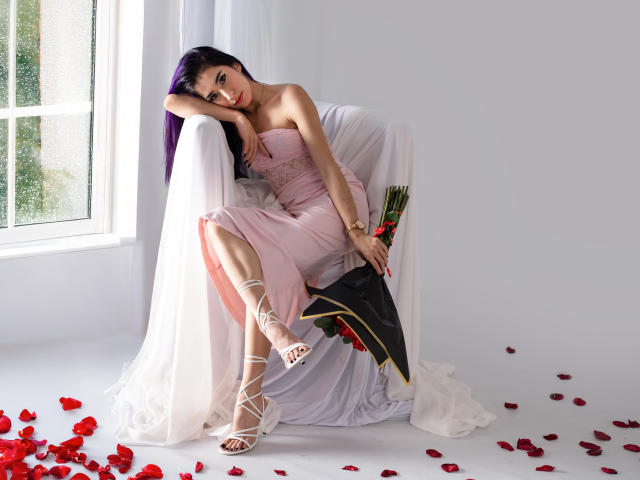 JuliaJobs with purple hair, pink dress, and white strappy heels, sitting in a white chair, holding roses, with red petals on the floor, and a black cape.