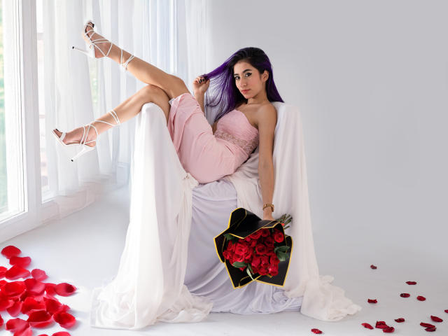 JuliaJobs with purple hair, pink dress, and white heels, legs up, holding red rose bouquet, white draped chair, rose petals on floor, looking at camera, sexy.