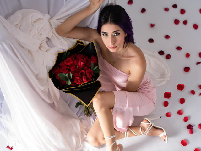 JuliaJobs with purple hair, pink dress, white heels, holding red rose bouquet, sitting on white fabric, surrounded by red rose petals, looking up with a slight smile.