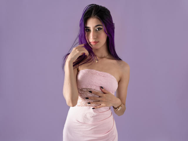 JuliaJobs with purple hair, wearing a pink lace strapless dress, touching her chin with one hand, holding her tits with the other, against a purple background.