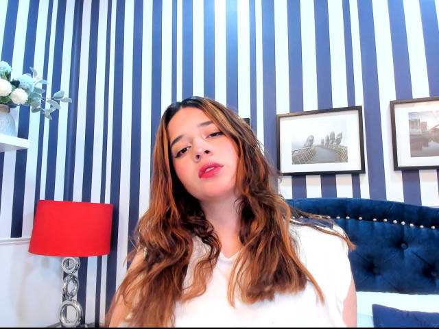 DaniVelasquez has wavy brown hair, red lipstick, and is wearing a white shirt. She's standing in front of a blue and white striped wall with a red lamp.