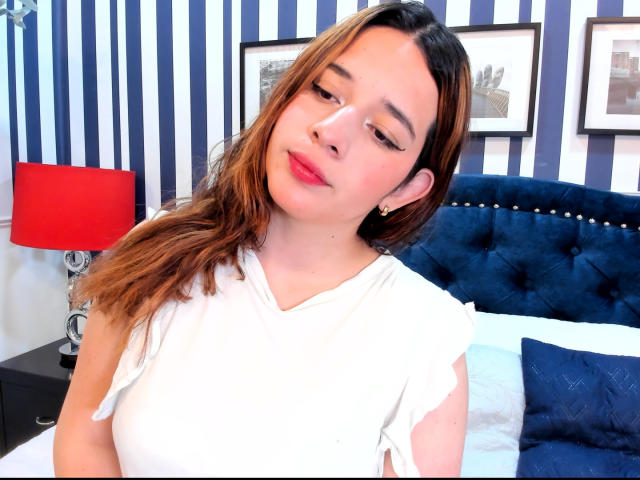 DaniVelasquez has medium tits, brown wavy hair, red lipstick, white shirt, and a blue-tufted headboard in the background. She's looking down with a slight smile.