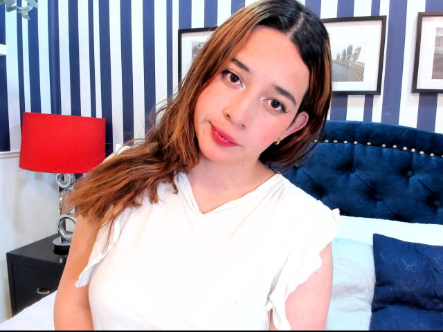 DaniVelasquez has brown hair, light skin, and red lipstick. She's wearing a white shirt, looking at the camera with a slight smile, and has small, cute tits.