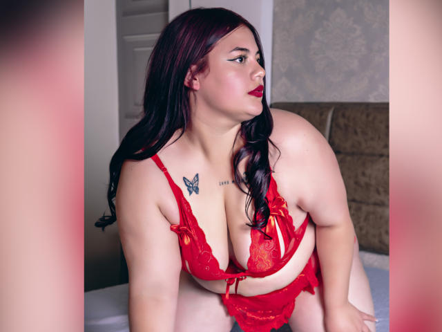 EllaRider with red lace lingerie, large tits, and a butterfly tattoo on her chest, leaning forward, dark red lipstick, black hair, looking to the side, in a bedroom.