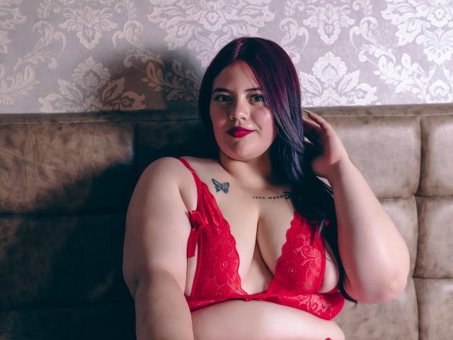 EllaRider met donkerrood haar, een rode kantbeha en een vlindertatoeage op haar borst, zittend op een bruine bank, hand in haar haar, lachend, waarbij haar grote borsten zichtbaar zijn..