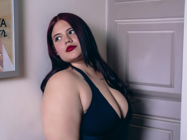 EllaRider has big tits in a black halter top, red lipstick, and long dark red hair, standing near a door with a slight smile. Her skin is fair.