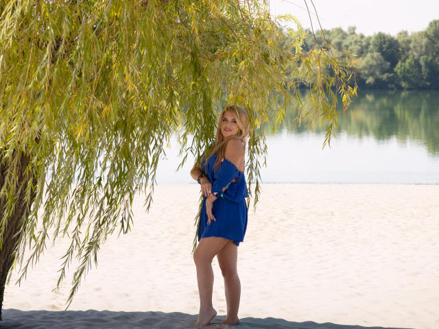 JulyaBailly stands under a weeping willow, wearing a blue dress, her ass slightly visible, blonde hair falling, smiling, barefoot on sandy shore, water and trees in background.