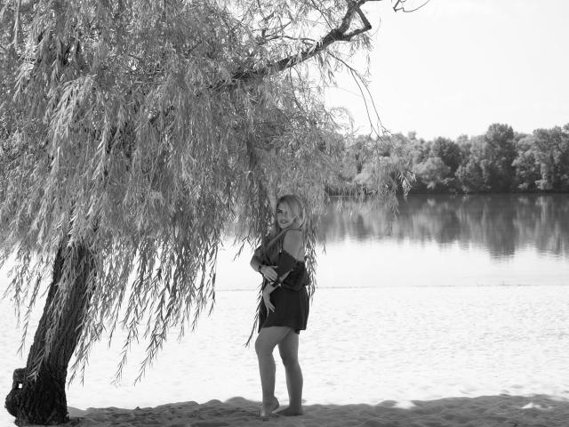 Black-and-white photo of JulyaBailly with long blonde hair, standing under a weeping willow, wearing a dark off-shoulder dress, by a calm lake, holding a small object.