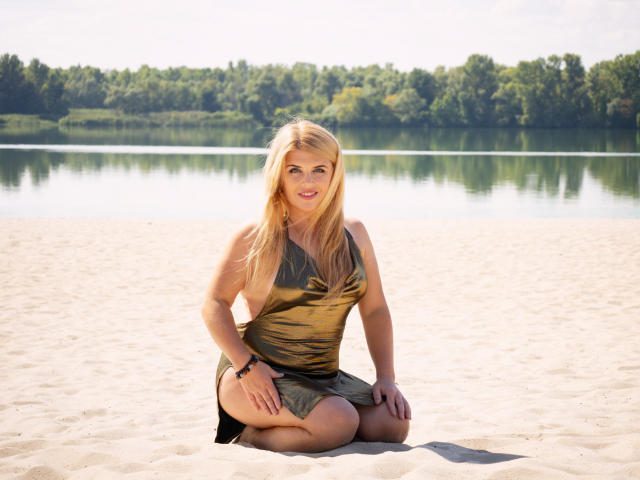 JulyaBailly, rubia, con pechos de tamaño medio, arrodillada en la arena, vestida con un vestido dorado, sonriendo a la cámara, con las manos en las rodillas, un lago y árboles en el fondo, un día soleado, con una pulsera en la muñeca derecha..