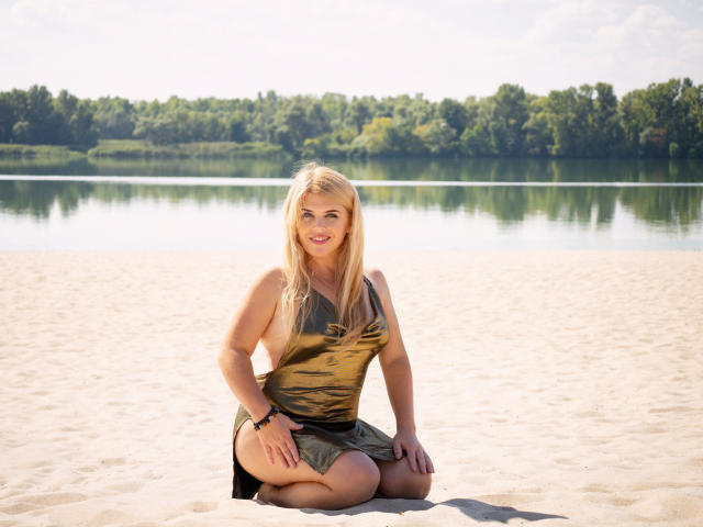 JulyaBailly, blonde with medium tits, wearing a green and gold patterned dress, kneeling on sandy shore, smiling, sunlight, green tree line in background, black bracelet on right wrist.