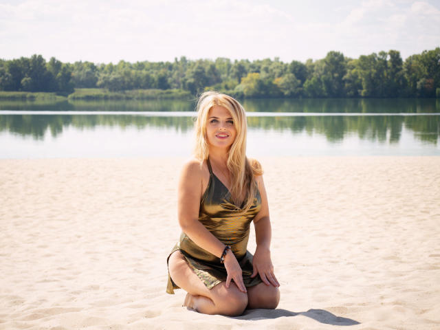 JulyaBailly, rubia, con senos, vestida con un vestido verde-marrón, arrodillada en una playa arenosa, sonriendo, con las manos en las rodillas, con una línea de árboles verdes y agua tranquila al fondo, un día soleado y brillante..