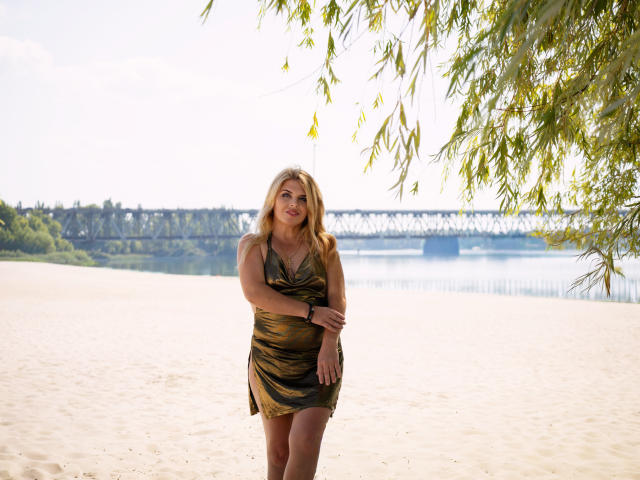 Blonde JulyaBailly with medium tits in a green and gold dress, standing on a sandy beach, leaning against a leafy tree, smiling at the camera.