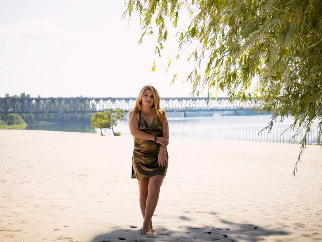 JulyaBailly está de pie en una playa arenosa, vestida con un vestido verde y negro a rayas de cebra, con cabello rubio, brazos cruzados y una ligera sonrisa, debajo de un árbol frondoso..