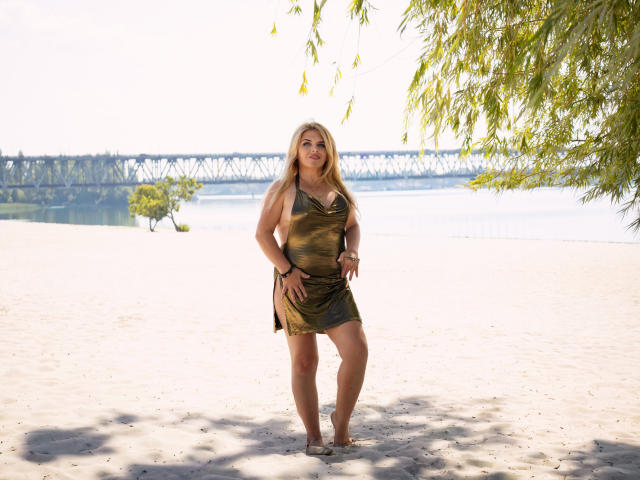 JulyaBailly está descalça em uma praia arenosa, vestindo um vestido verde, de estilo camuflado, com o cabelo loiro solto, as mãos na cintura, e uma ponte e água ao fundo..