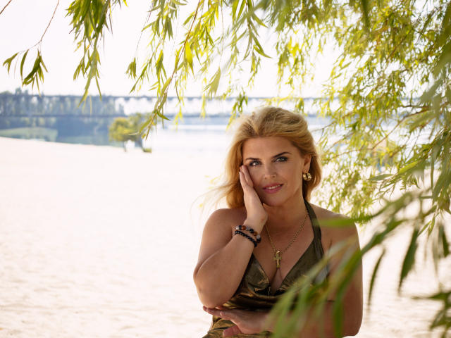JulyaBailly with blonde hair, green eyes, and a sexy smile, touching her cheek, wearing a green halter top, under leafy branches, with her hand on her ass.