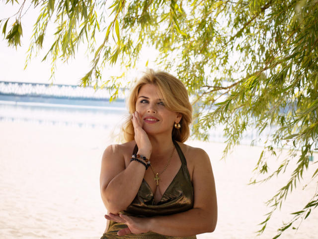 JulyaBailly with blonde hair, green halter top, hand on cheek, smiling, earrings, bracelets, under leafy branches, water and bridge in background, showing her tits and cute face.