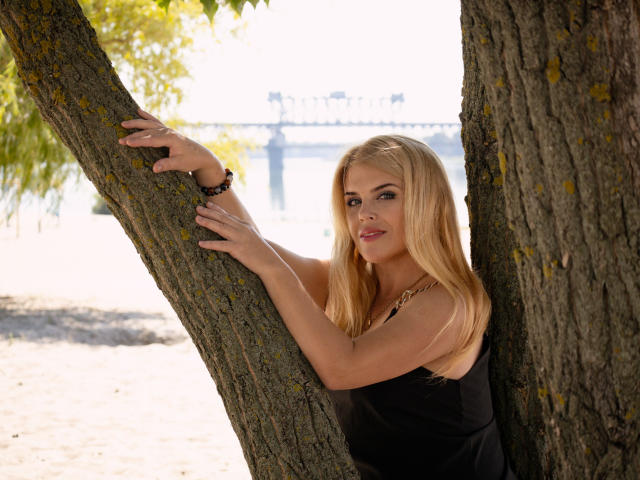 Blond JulyaBailly with long hair, black sleeveless top, and bracelet, leans against tree, holding it with both hands, showing her tits slightly. Watermark in bottom right.