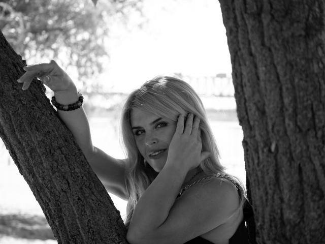 Black-and-white photo of JulyaBailly, blonde, leaning against tree, hands on face, bracelet on wrist, looking seductively at camera, tree trunks on both sides, water in background.