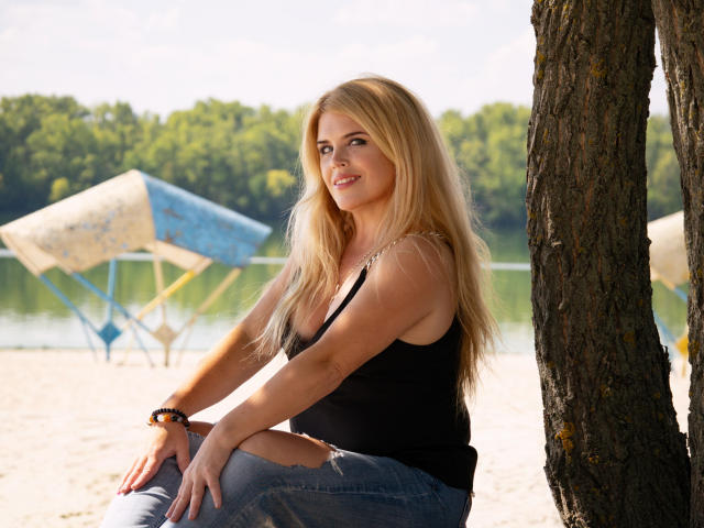 JulyaBailly with long blonde hair, black tank top, and ripped jeans, sitting against a tree, blue and white umbrella in background, wearing a black bracelet.