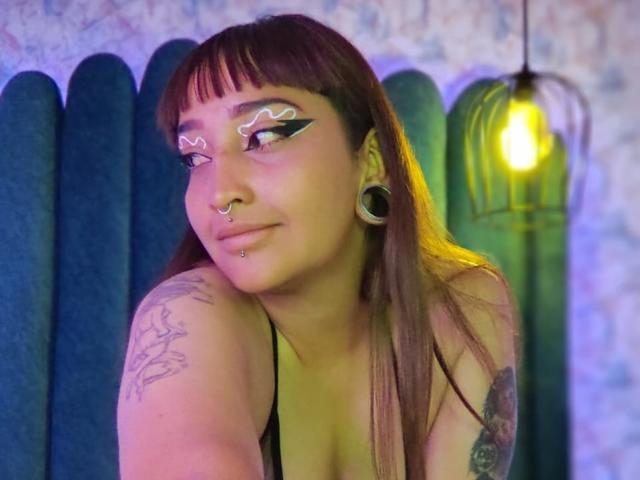 LadesIire with dark brown hair, nose ring, lip piercing, black eyeliner, tattoos, and large ear gauges, wearing a black top, sitting in front of a green velvet panel.