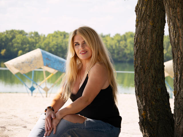 JulyaBailly, loira com cabelos longos, camiseta preta e calça jeans azul, está agachada em uma praia arenosa, sob uma árvore, parecendo feliz, com o bumbum e os seios levemente visíveis..