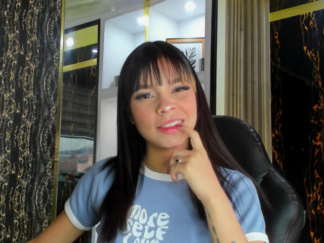 LunaFreya with black hair, light skin, blue shirt, finger touching her pink lips, sitting in black chair, looking at camera, ring on right finger, reflective windows behind.