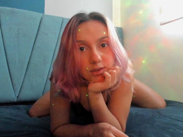 AlterEgoSecond con i capelli di colore rosa, sdraiata su un letto blu, con la testa appoggiata alla mano, che guarda la fotocamera, scintille nell'aria, carina, naso piccolo, labbra leggermente dischiuse, top nero..