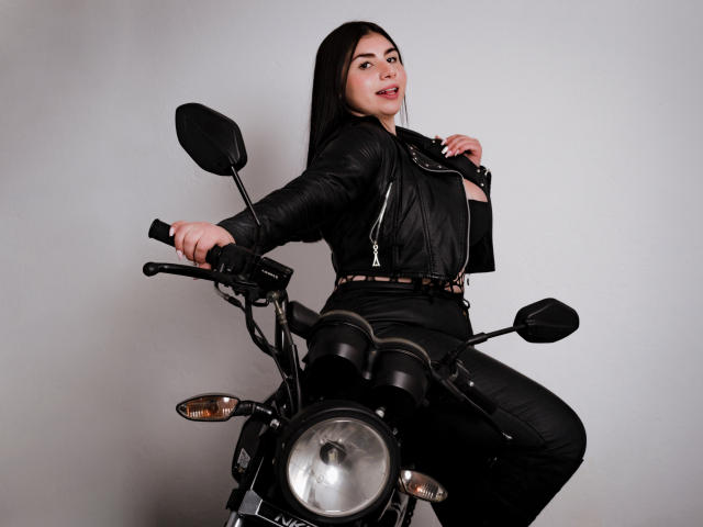 VenusxMusa with long black hair, wearing a black leather jacket and pants, sitting on a motorcycle, hands on the handlebars, looking over her shoulder, smiling.