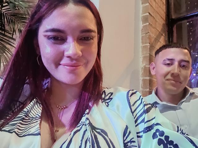 ThiagoJulieta with shiny face, red hair, wearing a zebra-patterned top, gold necklace, and hoop earrings, smiling at the camera, next to a man with a short haircut.