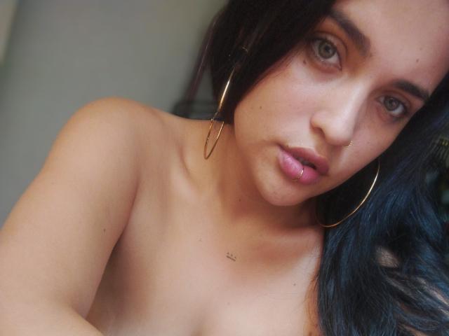 AgathaK has tan skin, black wavy hair, green eyes, and gold hoop earrings. She's topless, showing small tits, with a nose ring and slightly parted pink lips.