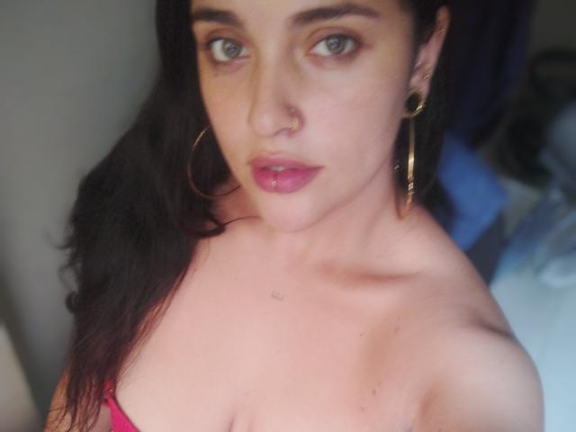 AgathaK with pink lips, hoop earrings, and dark hair, looking at the camera, showing her bare shoulders and small nose piercing, with a slightly blurred background.