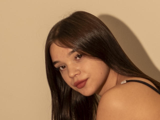 TiffanySavelin has long brown hair, brown eyes, and red lips. She's looking over her shoulder with a slight smile, wearing a black strap. Her ass is partially visible.