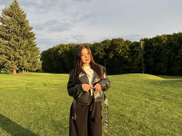 TiffanySavelin standing in a grassy field, wearing a black leather jacket, white turtleneck, and black pants, with long dark hair and a cute smile. Her ass is covered.