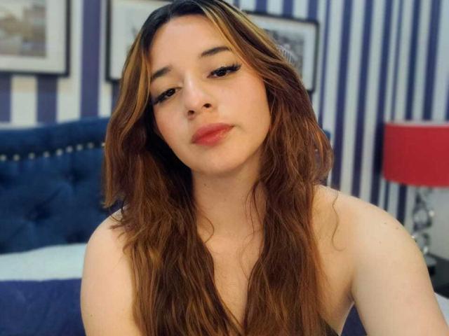 DaniVelasquez has long brown hair, a cute face, and is looking at the camera with a slight smile. She's wearing no clothes, showing her bare shoulders. Bedroom setting.
