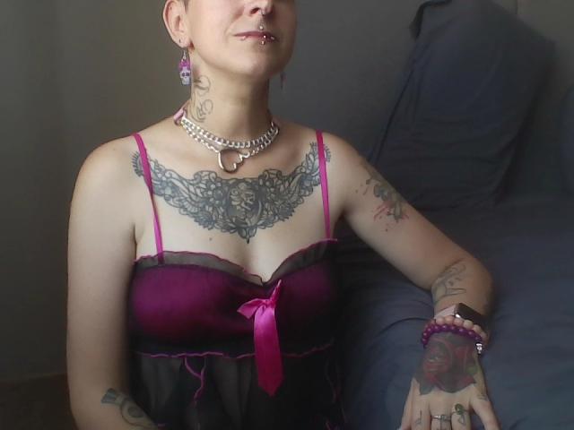 MissLilis with tattoos, pink lingerie, and a pink ribbon, wearing a necklace and earrings, showing her pierced lip and tattoos, sitting on a bed with gray pillows.