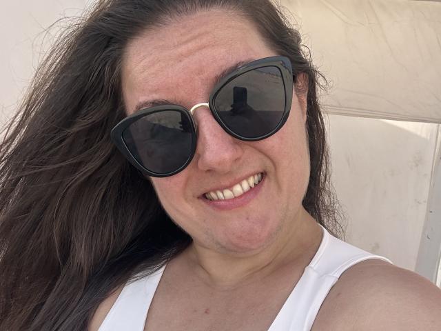 BadAngel with long brown hair, wearing black sunglasses, and a white tank top, smiling at the camera, showing off their teeth. Their face has slight freckles and a light skin tone.