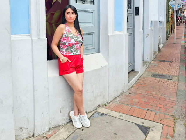 JainaRogers stands with hands in red shorts, wearing a floral top, white sneakers, and a neutral expression. Her ass is visible, and she has long black hair.